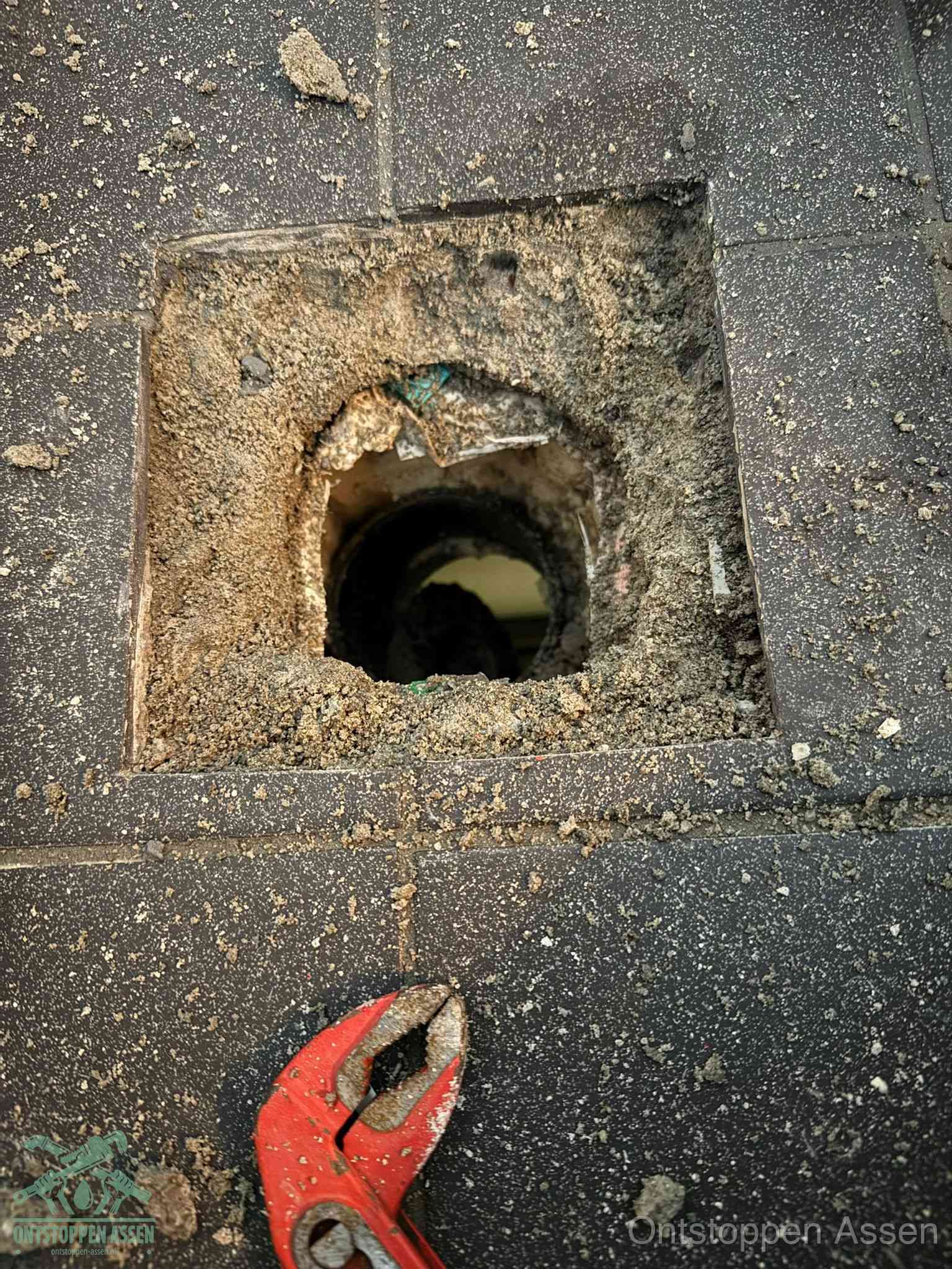 Een geopend rioolputje in een betontegelvloer met losgemaakt rooster en een zichtbare ronde afvoerbuis, het achterhalen waar de afvoerverstopping zit in Assen