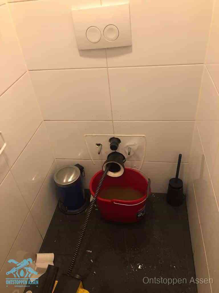 Een ontstopte wand-wc met verwijderde toiletpot waar een ontstoppingsveer in de wanduitgang verdwijnt, het achterhalen waar de verstopping zit in Assen