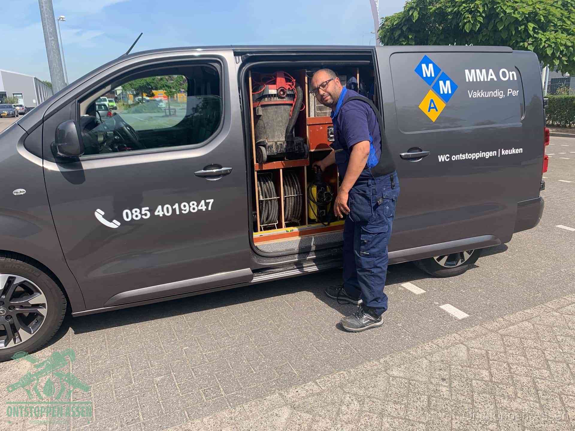 Een monteur bij de open schuifdeur van zijn bedrijfsbus op een parkeerplaats, klaar om de oorzaak van terugkerende afvoerverstoppingen aan te pakken in Assen