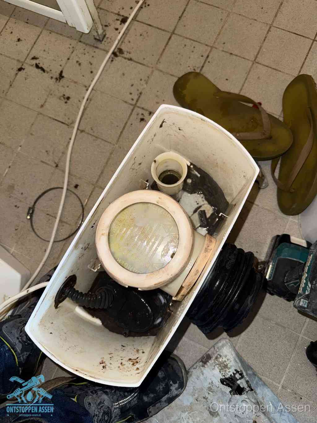 Een gedemonteerde toiletpot op de vloer met ontstoppingsveer en gereedschap ernaast, het verwijderen van een vastzittend voorwerp uit de wc in Assen