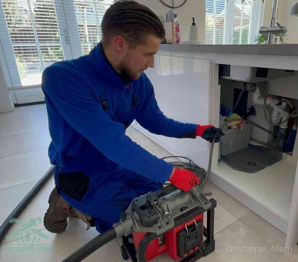 Een monteur met rode handschoenen knielt in een keuken en voert een elektrische ontstoppingsveer in tijdens het professioneel afvoer ontstoppen in Assen
